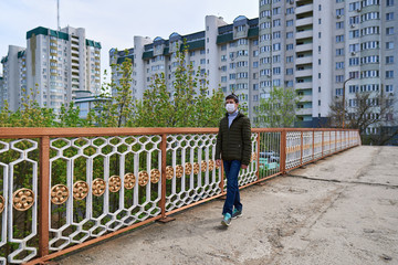 Fototapeta na wymiar teen boy walks down the street during the day, a pedestrian walkway and high-rise buildings with apartments, a residential area, a medical mask on his face protects against viruses and dust