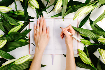 Feminine hands writing in notebook surrounded with green fresh flowers on white background. Trendy beautiful workspace with laptop and flowers in flat lay style