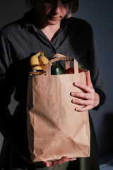 A young woman holds a paper bag with groceries - fruits and vegetables and looks into it. Vertical photo on a dark background