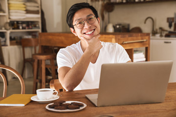 Image of asian man smiling and using laptop while sitting at table