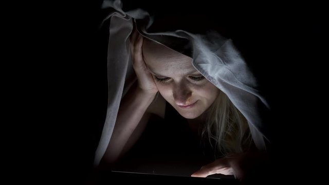 Woman suffering from insomnia uses a smartphone while lying in bed in the dark under covers.