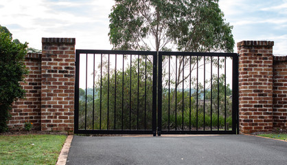 Black metal wrought iron driveway property entrance gates  set in brick fence, lights, green grass, garden trees