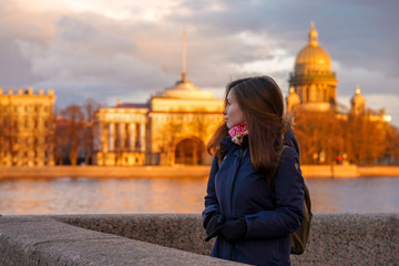 A brunette girl with long hair in a jacket and scarf stands on the embankment of St. Petersburg at sunset with a view of St. Isaac's Cathedral and the Golden buildings