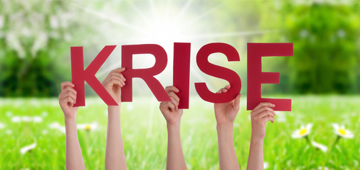 People Hands Holding Colorful German Word Krise Means Crisis. Sunny Green Grass Meadow As Background