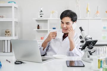 Obraz na płótnie Canvas A male scientist with black hair wearing white coat using a mobile phone device sipping coffee with a laptop and a microscope in a laboratory setting with test tubes. Looking relax during break time.