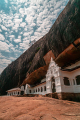 Biała buddyjska świątynia w Dambulla z posągiem Buddy oraz stupą na tle niebieskiego nieba, mieszcząca się pod skałą, Sri Lanka.