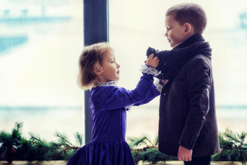 Two kids, boy and girl in vintage retro clothes near window sill. Girl in vintage violet dress touching boys big brown scarf on neck. Valentine day. Love. Together. Retro post card with children