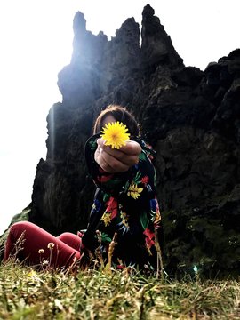 Woman Holding Yellow Dandelion While Sitting On Grassy Field Against Rocky Mountains