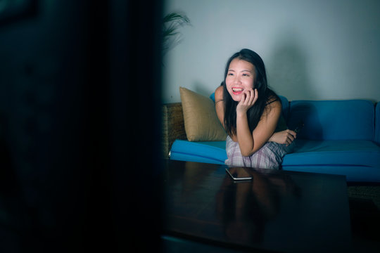 young happy and beautiful Asian Korean woman watching TV  at living room sofa couch enjoying funny television Korean drama show or comedy movie relaxed and cozy