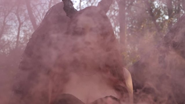 Brunette girl maleficent with wings and horns walking in the forest through the smoke