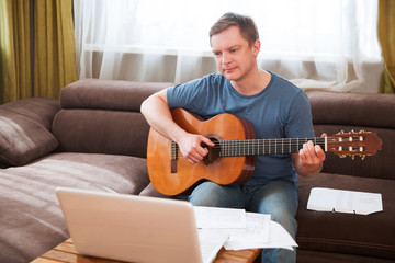 Man playing acoustic guitar and watching online lesson on laptop while practicing at home. Stay home. quarantine. Online training, online classes.
