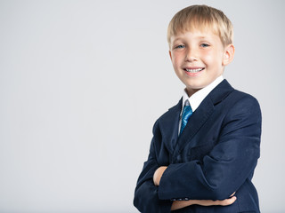 Portrait of white smiling  kid in a blue business suit looking at camera.  Photo of a happy businessman boy.  Positive blond boy dressed in a formal suit with tie with with crossed arms on breast