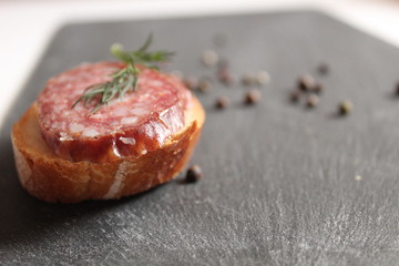 still life of food sandwich of white bread and smoked sausage on a gray black stone table next to the pepper black peas and seasoning herbs dill