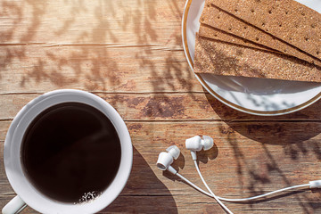 A cup of coffee, a saucer with crisp bread, headphones on a wooden tabletop. Top view.  Copy space.