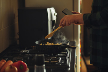 Young man with beard cooking dinner and using his smart phone at the kitchen.