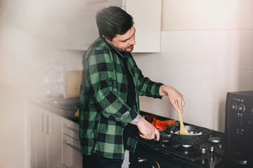 Young man with beard cooking dinner and using his smart phone at the kitchen.