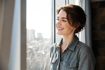 Image of beautiful young joyful woman smiling and looking at window