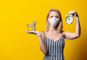 Blonde woman in face mask and gloves holds shopping cart and alarm clock on yellow background