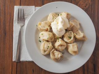 Traditional russian large dumplings with sour cream on a white plate and wooden table, and a metal fork.