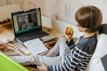 Distance learning for children during the coronavirus epidemic. The boy sits on the bed and receives a school assignment using the Internet and a laptop.