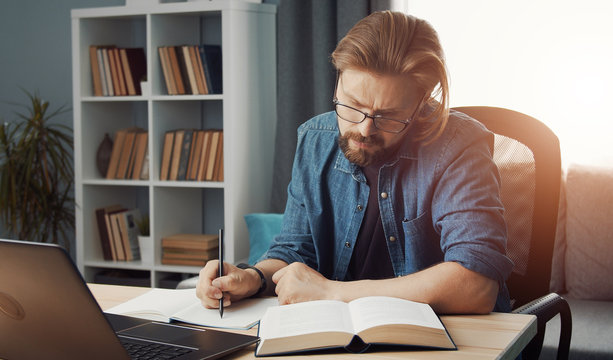Casual man working from home looking at book sitting in front of laptop at table in living room