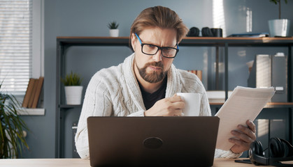 Busy adult male looking at laptop screen holding papers and cup working from home office