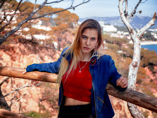 pretty young woman looking at the camera with blue eyes and blond hair wears a denim sweater and a red shirt, leaning against a wooden railing in the background the coast is seen