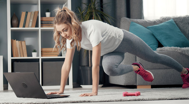 Girl watching online fitness session on laptop doing high plank with crunches staying in flat