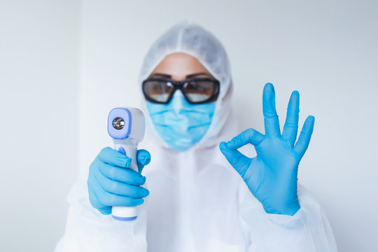 Doctor holding infrared thermometer and showing ok gesture. Portrait of medical professional in protective clothing measuring contactless fever at Covid-19 test center during coronavirus epidemic.