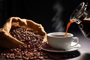 Pouring coffee with smoke on a cup and coffee beans on burlap sack on black background.