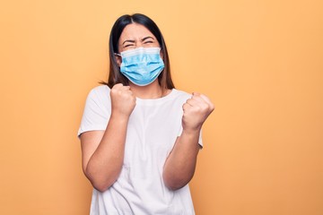 Young woman wearing protection mask for coronavirus disease over yellow background celebrating surprised and amazed for success with arms raised and eyes closed