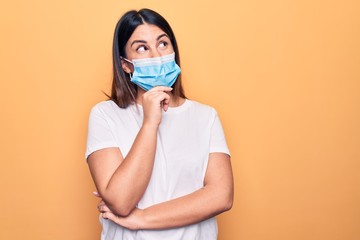 Young woman wearing protection mask for coronavirus disease over yellow background thinking...