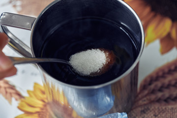A teaspoon with sugar is lowered into an iron mug with tea, coffee. Close-up.