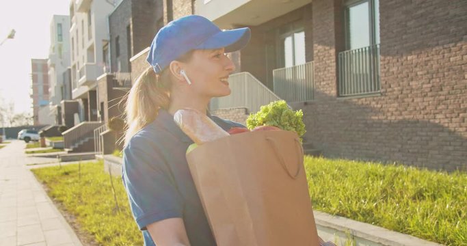Female young Caucasian delivery courier bringing fresh food from market to senior retired man and handing packet outdoor. Woman carrying bag with grocery when working in delivering.