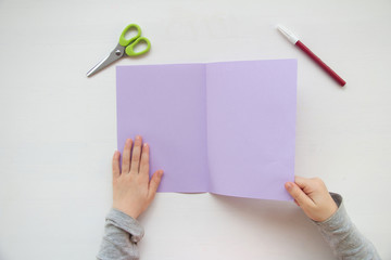 Child made a greeting card. Step 3. Child holds a card in his hands. Tools and materials for children's art creativity on table. Mother's day or March 8 greeting card DIY idea. 