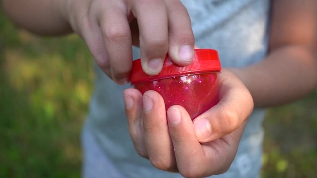Closeup view video footage of two kids hands holding pink transparent slime in transparent plastic box covered with red cap. Boy going to play slime glue queezing and pulling it. 