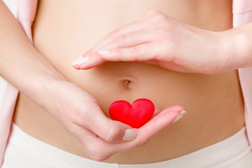 Obraz na płótnie Canvas Young woman hand holding red bright heart on naked belly. Emotional loving moment in pregnancy time. Baby expectation. Love, happiness and safety concept. Closeup. Front view.