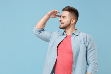 Smiling young bearded guy 20s in casual shirt posing isolated on pastel blue background studio portrait. People lifestyle concept. Mock up copy space. Hold hand at forehead looking far away distance.