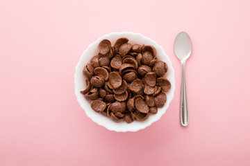 White bowl of chocolate corn flakes with spoon on light pink table background. Pastel color. Simple daily healthy breakfast. Closeup. Top down view.