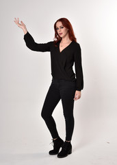 Portrait of a pretty girl with red hair wearing black jeans, boots and a blouse.  full length standing pose, facing the camera with hand gestures on a studio background.