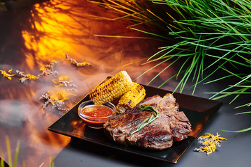 Juicy T-Bone steak with grilled corn and barbecue sauce on a black square plate on a dark smoky background with warm lighting. Background decorated with beautiful greenery. Close-up. Space