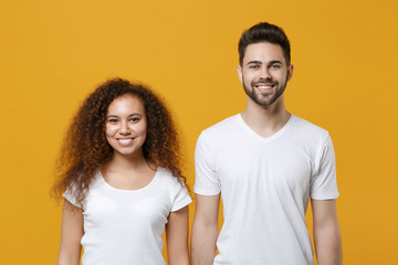 Smiling young couple two friends european guy african american girl in white t-shirts posing isolated on yellow wall background in studio. People lifestyle concept. Mock up copy space. Looking camera.