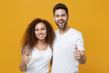 Cheerful young couple two friends european guy african american girl in white t-shirts posing isolated on yellow background. People lifestyle concept. Mock up copy space. Hugging, showing thumbs up.