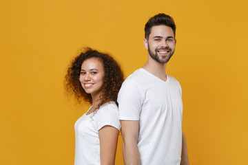 Smiling young couple friends european guy african american girl in white t-shirts posing isolated on yellow background in studio. People lifestyle concept. Mock up copy space. Standing back to back.