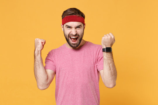 Screaming young bearded fitness sporty guy sportsman in headband t-shirt in home gym isolated on yellow background. Workout sport motivation concept. Wearing smart watch on hand, doing winner gesture.