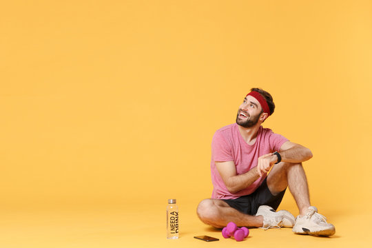 Funny Fitness Sporty Guy Sportsman In Headband T-shirt In Home Gym Isolated On Yellow Background. Workout Sport Motivation Concept. Sit With Smart Watch On Hand, Water Bottle, Mobile Phone, Dumbbells.