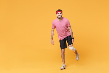 Fototapeta na wymiar Ingured dissatisfied young bearded fitness guy 20s sportsman in headband t-shirt in home gym isolated on yellow background. Workout sport motivation concept. Touching leg with elastic bandage on knee.
