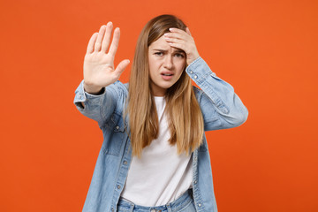 Worried young woman girl in casual denim clothes posing isolated on orange background studio portrait. People lifestyle concept. Mock up copy space. Showing stop gesture with palm, put hand on head.