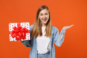 Funny young woman girl in casual denim clothes isolated on orange background. Valentine's Day, Women's Day, birthday, holiday concept. Hold red present box with gift ribbon bow, pointing hand aside.