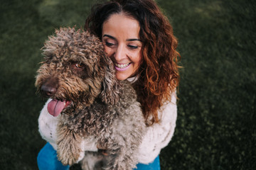 A beautiful woman is in the meadow with her dog. The owner is hugging her pet while looking at it with love. They are enjoying a day in the park. The pet is a Spanish water dog with brown fur.